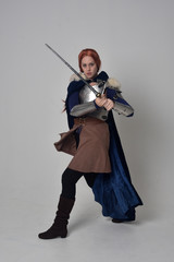 full length portrait of a  red haired girl wearing medieval warrior costume and steel armour and a fur cloak, standing pose on grey studio background.