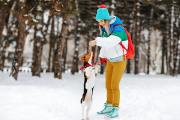 Fototapeta na wymiar Portrait of cheerful young woman in warm stylish clothes and bagpack playing with her beagle dog in winter park. Friendship, pet and human.