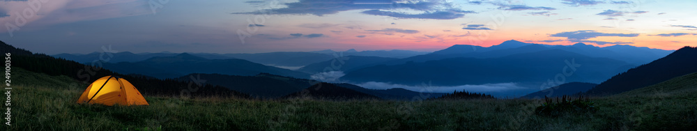 Wall mural orange tourist tent illuminated from inside stands in mountains above clouds