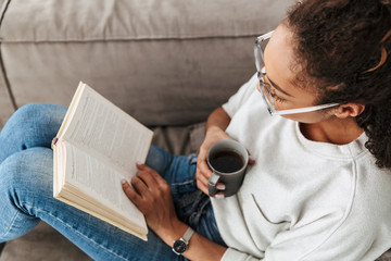 Image of brunette african american girl reading book and drinking tea, while sitting on sofa in...