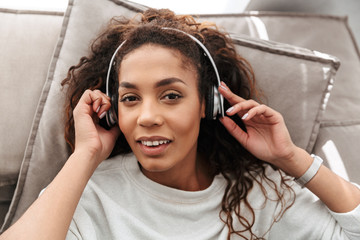 Photo of attractive african american woman listening to music using wireless headphones, while lying on sofa at home