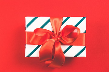 Beatiful gift with red satin bow and covered in a white paper with green line on a red background. Festive concept. Top view.