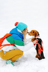 Fototapeta na wymiar Portrait of pretty young woman in warm stylish clothes and bagpack kiss with her beagle dog in winter park. Friendship, pet and human.