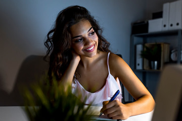 Young woman working and studying late at night, she is connecting with her computer and writing down notes on a notepad