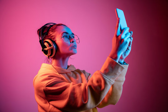 Fashion pretty woman with headphones listening to music and making selfie photo over red neon background at studio.