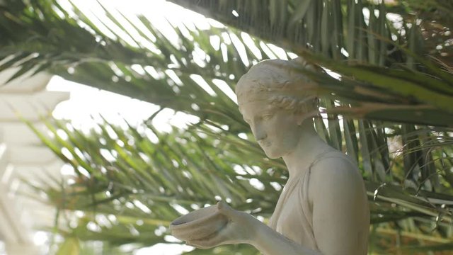 Slow motion track around a marble statue of a woman with palm leaves in the background
