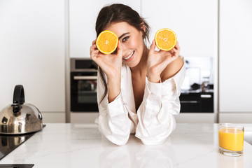 Image of european woman 20s wearing silk leisure clothing, holding two pieces of orange in kitchen