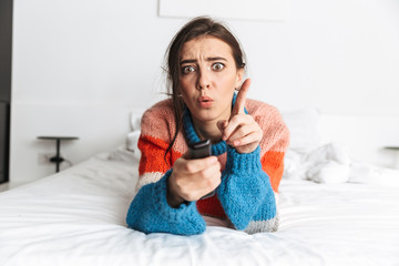 Portrait of cheerful woman 20s holding remote control, while lying in bedroom