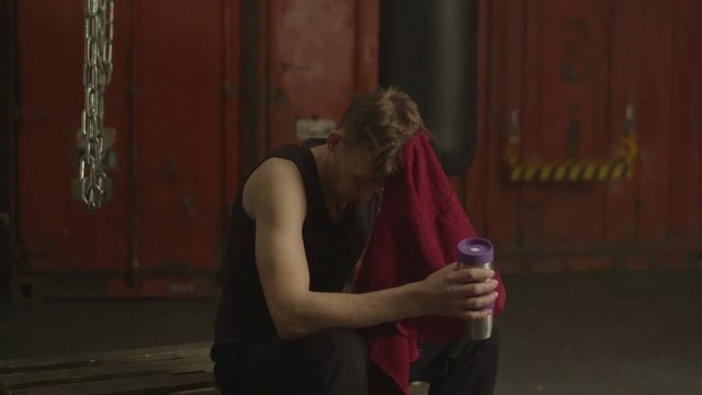 Exhausted athletic man wiping sweat with towel and drinking energy drink after powerful workout while sitting on wooden pallet in rustic gym. Active fit guy taking a break from exercising in gym.