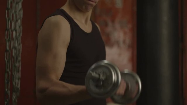 Midsection of athletic muscular man doing heavy dumbbell exercise for biceps in gym. Hand of spoty fit guy lifting dumbbell while working out weight training and bodybuillding in back-block gym.