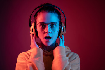 Surprised pretty woman with headphones listening to music over red neon background at studio.