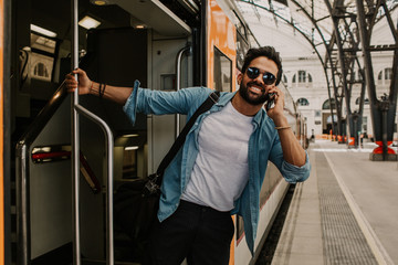 Confident Attractive young hispanic wearing sunglasses and blue shirt male holding smartphone hand and calling to friends while he standing at the train wagon on platform.