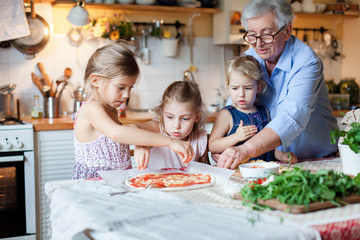 Children chefs are cooking italian pizza in cozy home kitchen for family dinner. Grandmother is teaching cute kids to cook food. Three little girls are helping old senior woman. Lifestyle moments.