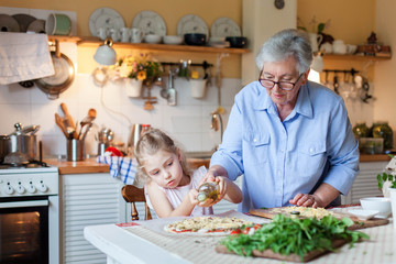 Kid is helping grandmother to cook italian pizza in cozy home kitchen for family dinner. Cute girl is cooking homemade food. Old senior woman is teaching child. Children chef concept.