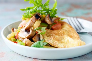 Homemade omelette with mushrooms, vegetables and arugula on a plate. Wonderful healthy breakfast near the window with a cup of coffee.