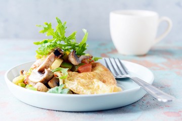 Homemade omelette with mushrooms, vegetables and arugula on a plate. Wonderful healthy breakfast near the window with a cup of coffee.