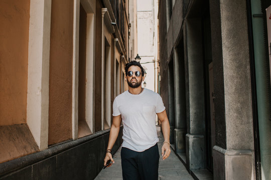 Content Attractive Hispanic Man In White Tshirt Walking Old Town Streets Holding Mobile Phone Hand.Bearded Hipster Male Enjoying City Travel On Gotic District Streets