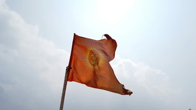 A saffron colored Hindu flag flying during daytime with the sky and clouds in the background. The work 'Om' is written in Hindi language.