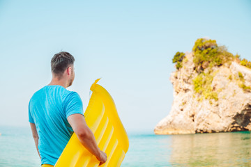man standing with yellow inflatable mattress and looking at sea with rocky cliff