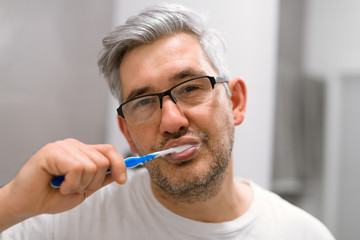 middle aged gray haired man brushing teeth in bathroom
