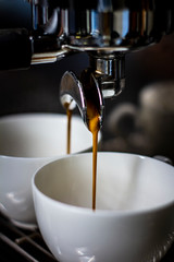 Two white cups standing on the grating of coffee machine and coffee pouring into them