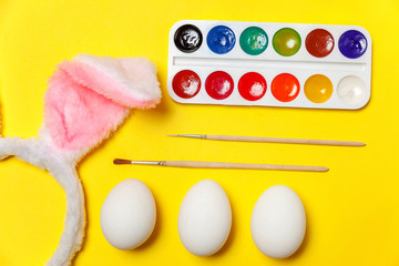 Happy Easter concept. Preparation for holiday. Decorative eggs colorful paints and bunny ears furry costume toy isolated on trendy yellow background. Simple minimalism flat lay top view copy space