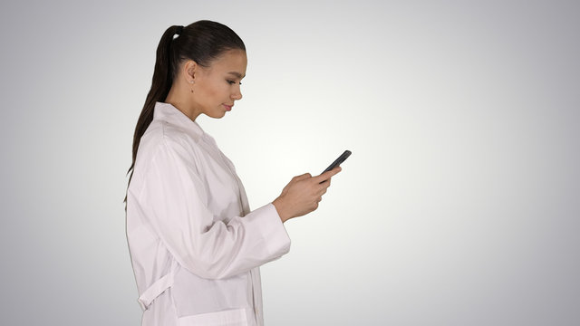 Cheerful cute young woman doctor typing text on phone while walking on gradient background.