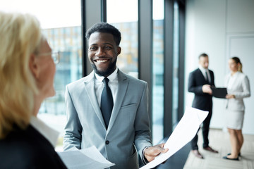 Young smiling African-american businessman in formalwear showing new contract to his colleague in...
