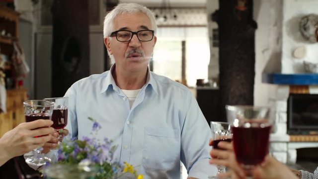 Panning of aged man pronouncing some words as toast, clinking glasses with other members of family sitting at dinner table