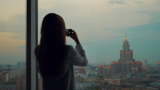 Young beautiful woman or tourist taking photograph with mobile cellphone smartphone device of skyline in Moscow Russia view from her apartment. Girl to share pic on social media photo site.