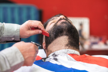 Barber shaving the beard of a handsome bearded man with an electric razor at the barber shop .