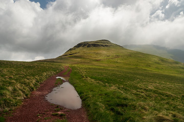 Lonely red road with rain puddles on beautiful mountain with green grass and clouds covering the peak. Beautiful landscape on Stara Planina mountain in Serbia.