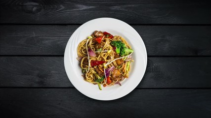 Rotating udon stir fry noodles with meat or chicken and vegetables in a white plate on black wooden background. Top view
