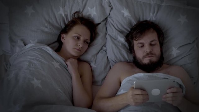 Caucasian Couple Fighting, arguing on Bed. Bearded Man addicted to gadget - Smart Phone, ignoring wife flirting in bed. Offended Woman lying turned back to her Husband at night.