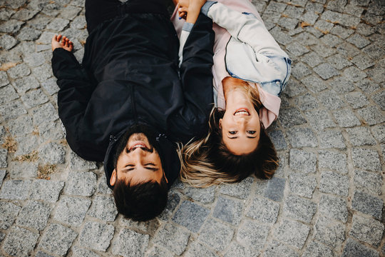 Lovely young caucasian couple having fun laughing and holding by hands while lying on the ground in the city.