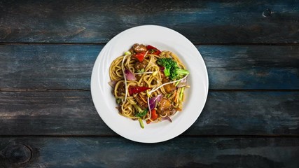 Rotating udon stir fry noodles with meat or chicken and vegetables in a white plate on wooden background. Top view