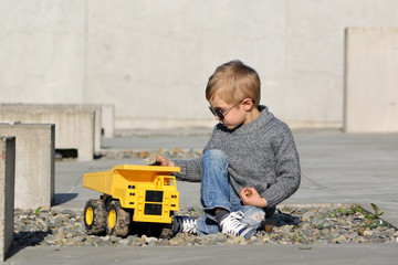 A four years old smiling boy in sunglasses playing a yellow track on concrete background.