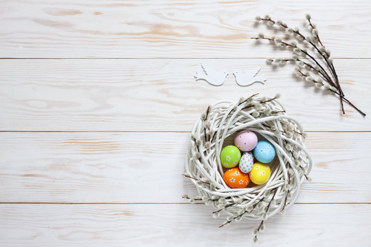 Easter decoration – nest with colorful decorative eggs, dove figurines and pussy willow branches. Top view, close up, flat lay on white wooden background