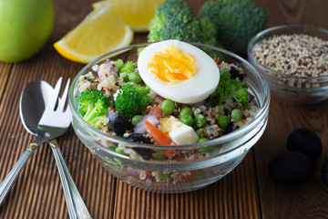 Bowl with quinoa and different vegetables - egg, broccoli, tomato, green peas, red onions. Wooden table. Healthy food.