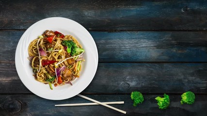 Rotating udon stir fry noodles with meat or chicken and vegetables. Decorated with fresh broccoli and chopsticks in a white plate on wooden background. Top view with the copy space for your text