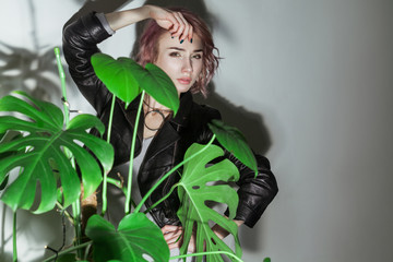 beautiful woman with makeup and short pink hair in grey dress and black leather jacket posing near green palm leaves on grey background. indoor studio shot.
