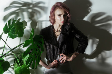 beautiful woman with makeup and short pink hair in grey dress and black leather jacket posing near green palm leaves on grey background. indoor studio shot.