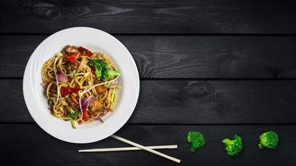Rotating udon stir fry noodles with meat or chicken and vegetables. Decorated with fresh broccoli and chopsticks in a white plate on black wooden background. Top view with the copy space for your text