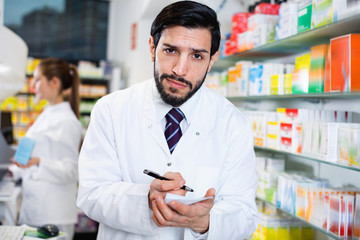 Male specialist is attentively stocktaking medicines with notebook near shelves in pharmacy.
