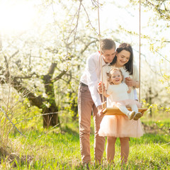 Cheerful family shakes a child on a swing, in a flowered garden or park. Warmly, love, spring and summer mood
