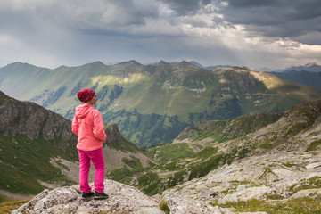 Obraz premium view from the back on girl in pink pants standing on a cliff hands in pockets on a mountain background under storm sky