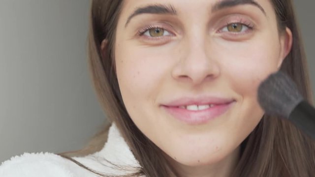 Girl just woke up, dressed in white bath gown, doing mornig light makeup, applying some powder with brush, smiling a lot, showing her teeth, looking at camera with big beautiful eyes. Close up concept