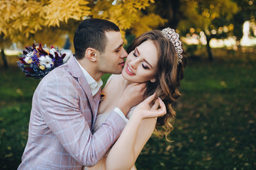 Fototapeta premium Newlyweds in love embrace in the fall in the park with yellow foliage. Stylish groom in a plaid jacket kisses a pretty bride in a beige dress with a crown. Wedding photography. Princess Bride