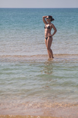 Fototapeta na wymiar brunette girl with a beautiful figure in a black bikini and sunglasses standing in the sea ankle-deep in the water on a sandy shoal