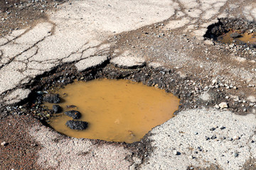 Potholes on a paved road. The road is in very poor condition. There is dirty water in the potholes.